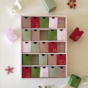 Puede incluir: Un calendario de adviento rectangular con veinticinco cajones individuales en tonos rojo, verde, rosa y beige, cada uno marcado con números dorados. Varios cajones sueltos y recortes florales de papel están esparcidos alrededor.