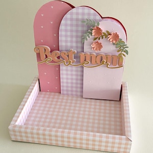 Puede incluir: Un expositor decorativo con una base de bandeja y tres paneles arqueados. Los paneles presentan cuadros vichy rosas, corazones rosas y un fondo rosa claro. Las palabras "Best mom" están en cursiva, con flores y hojas de papel.