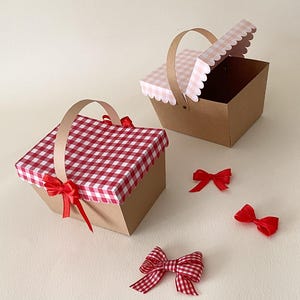Puede incluir: Dos cestas de picnic de papel marrón con tapas a cuadros rojos y blancos y asas color canela. Una cesta está cerrada con una cinta roja y la otra está abierta. Tres lazos rojos están esparcidos cerca.
