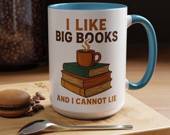 May include: White ceramic mug with a blue interior and handle. The mug features the text "I LIKE BIG BOOKS AND I CANNOT LIE" with an illustration of a coffee cup on top of a stack of books. A spoon and a macaron are on the wooden surface.
