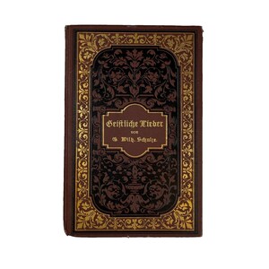 Pode incluir: Um livro retangular de capa dura com uma capa castanho-escura apresentando bordas de filigrana dourada e um painel central preto. O texto na capa diz Geistliche Lieder von G. Wilh. Schulze em letras douradas dentro de uma moldura.