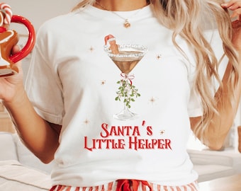May include: A white t-shirt with the text "Santa's Little Helper" in red, featuring a cocktail graphic with a gingerbread man. The person is also wearing red and white striped pajama pants and holding a gingerbread man mug.
