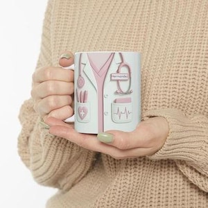 Könnte beinhalten: Eine weiße Keramik-Tasse mit einem Arztkittel-Design in Hellblau und Rosa. Das Design umfasst ein Stethoskop, Taschen und den Namen "Hernandez". Die Tasse wird in den Händen einer Person gehalten.