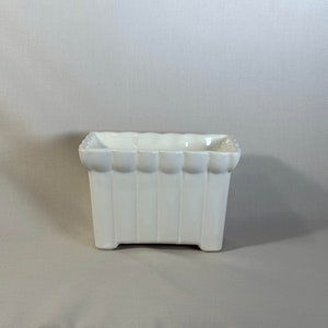 Peut inclure: Pot en céramique blanche de forme rectangulaire avec des bords festonnés. Le pot a des nervures verticales sur les côtés et une base légèrement surélevée.