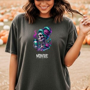 Puede incluir: Una camiseta gris carbón con un gráfico de un esqueleto con cabello verde azulado y rosas rosas sosteniendo una bebida, con el texto MOMBIE debajo. La camiseta se combina con una gorra de béisbol verde oliva con el texto PUMPKIN SPICE en letras blancas.