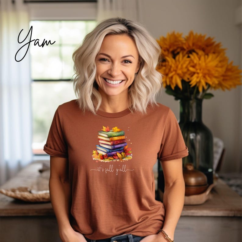 A rust-colored short-sleeve t-shirt featuring a graphic of a stack of colorful books surrounded by autumn leaves. Below the books, the text reads "it's fall y'all" in a white cursive script. The shirt has a crew neckline and hemmed sleeves.