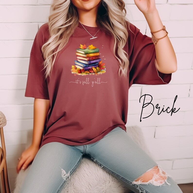 A brick red short-sleeve t-shirt featuring a graphic of a stack of colorful books surrounded by autumn leaves. Below the books, the text reads "it's fall y'all" in a white cursive script. The shirt has a crew neckline and a loose fit.