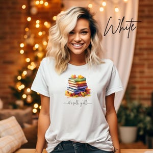 A white short-sleeve t-shirt featuring a graphic of a stack of colorful books surrounded by autumn leaves. Below the books, the text reads "it's fall y'all" in a cursive script. The word "White" is written in black script in the top right.