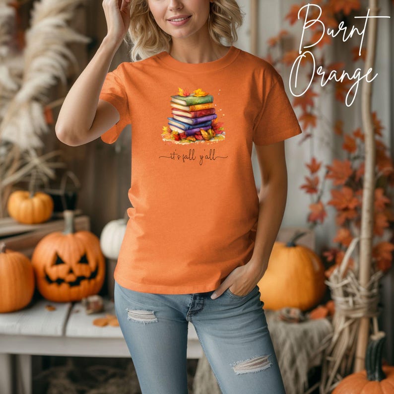 A burnt orange short-sleeve t-shirt featuring a graphic of a stack of colorful books surrounded by autumn leaves. Below the books, the text reads 'it's fall y'all' in a cursive script. The shirt has a crew neckline and a standard fit.