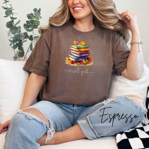 A brown short-sleeve t-shirt featuring a graphic of a stack of colorful books surrounded by autumn leaves. Below the books, the text reads "it's fall y'all" in a cursive script. The shirt has a crew neckline and a relaxed fit.