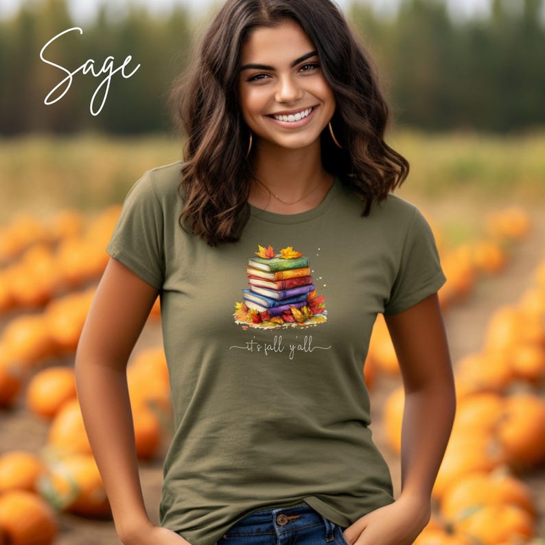 A sage green t-shirt featuring a graphic of a stack of colorful books surrounded by autumn leaves. Below the books, the text reads "it's fall y'all" in a white cursive script. The word "Sage" is printed in white in the top left corner.