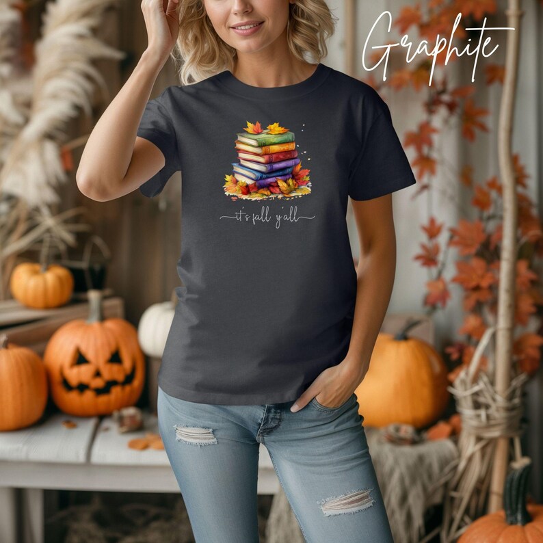 A dark gray t-shirt featuring a graphic of a stack of colorful books surrounded by autumn leaves. Below the books, the text reads "it's fall y'all". The shirt is paired with light blue denim jeans that have distressed knee details.