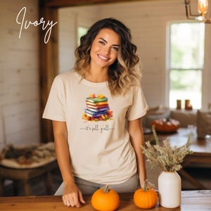 An ivory short-sleeve t-shirt featuring a graphic of a stack of colorful books surrounded by autumn leaves. Below the books, the text reads "it's fall y'all" in a cursive script. The shirt has a crew neckline and a relaxed fit.