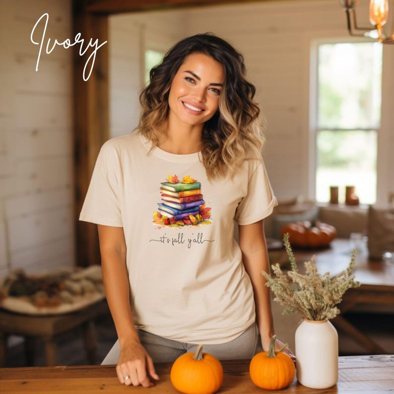 An ivory short-sleeve t-shirt featuring a graphic of a stack of colorful books surrounded by autumn leaves. Below the books, the text reads "it's fall y'all" in a cursive script. The shirt has a crew neckline and a relaxed fit.
