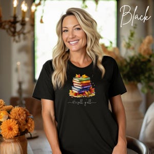 A black short-sleeved t-shirt featuring a graphic of a stack of colorful books surrounded by autumn leaves. Below the books, the text reads "it's fall y'all" in a white cursive script. The shirt has a crew neckline and a straight hem.