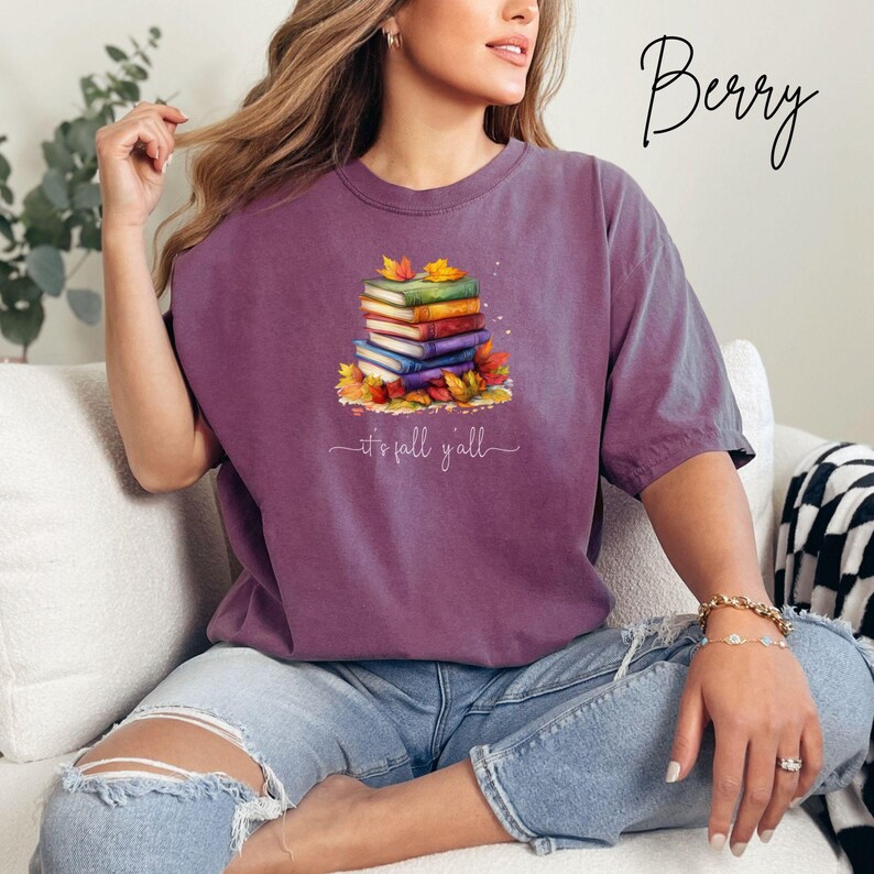 A berry-colored short-sleeve t-shirt featuring a graphic of a stack of colorful books surrounded by autumn leaves. Below the books, the text reads "it's fall y'all" in a white cursive script. The shirt has a crew neckline and a relaxed fit.