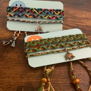 May include: Two sets of handmade friendship bracelets. One set features black, blue, yellow, and pink patterns. The other set has green and brown patterns. Each bracelet has a small elephant charm and decorative tassels.