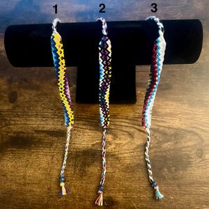 May include: Three handmade friendship bracelets with intricate woven patterns. The bracelets feature a variety of colors, including yellow, blue, purple, and red. Each bracelet has a beaded accent and a tassel at the end. The bracelets are displayed on a black velvet surface.