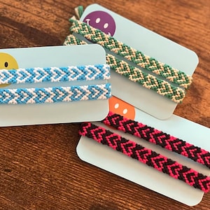 May include: Three pairs of friendship bracelets in blue and white, green and beige, and black and pink chevron patterns. Each pair is displayed on a light blue card with a smiley face or a simple graphic.