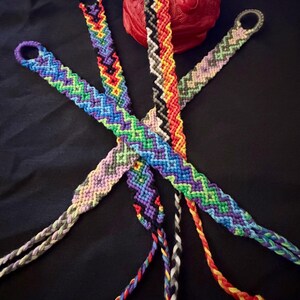 May include: A collection of colorful friendship bracelets with intricate woven patterns. The bracelets feature a variety of colors, including blue, green, purple, red, and yellow. A red, textured decorative sphere is also present. The bracelets are displayed against a black background.