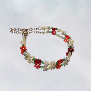 May include: A delicate bracelet made with small, colorful beads. The bracelet features a floral design with red, green, and white beads. The bracelet has a gold clasp.