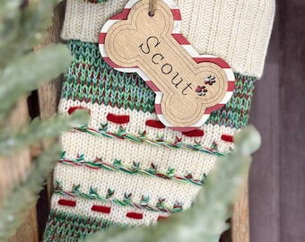 May include: A Christmas stocking with a cream-colored cuff and a patterned body in green, red, and white. A wooden bone-shaped tag with the name "Scout" and paw prints hangs from the stocking. A green and red pom-pom and a wooden bead are attached to the stocking.