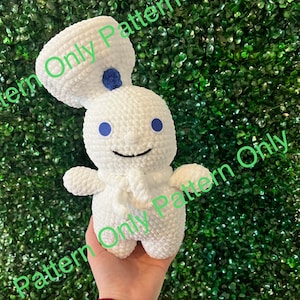 May include: White crocheted plush toy with blue eyes and a blue button on its head. The toy is smiling and has a white crocheted scarf. The toy is being held in a hand against a green background.