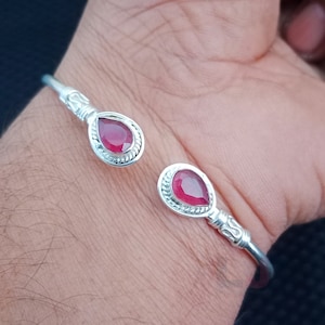 Puede incluir: Un brazalete de plata con dos acentos de piedras preciosas rojas. El brazalete está abierto y tiene un diseño decorativo.