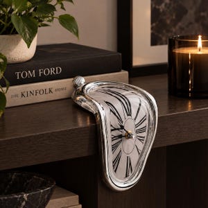 May include: A silver-toned melting clock with a white face and black Roman numerals. The clock features a curved, distorted frame designed to drape over the edge of a flat surface, with black hour and minute hands centered on the dial.