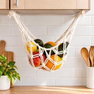 May include: A cream-colored macrame fruit hammock suspended between two wooden dowels under a kitchen cabinet, containing a variety of fresh produce including a red apple, a yellow lemon, an orange, and two dark green avocados.