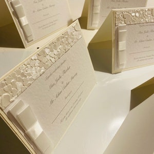 May include: White wedding invitation cards with a raised floral design and a white satin ribbon. The text on the card reads "Together with their families Miss Jolie Fletcher & Mr Adam Cashin-Murray Save the Date".