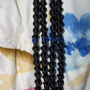 Peut inclure: Quatre porte-clés macramé noirs avec des fermoirs argentés. Chaque porte-clés présente un motif de corde torsadée avec des accents bleus. Les porte-clés sont présentés sur un tissu à motifs floraux.