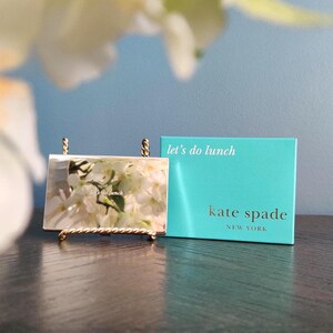 May include: A gold-toned metal picture frame with a floral image and the text "let's do lunch" is displayed on a small gold stand. A teal box with the text "let's do lunch" and "kate spade new york" is next to the frame.