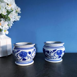 May include: Two small, white ceramic pots with blue floral designs. The pots have a rounded shape with a flared rim and a small base. The background is a solid blue color, and a vase of white flowers is visible.