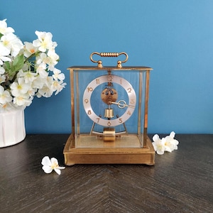 May include: A rectangular brass-framed clock with a clear glass enclosure. The clock face is white with black numerals and hands. A gold-colored handle is on top. The clock sits on a dark wooden surface.