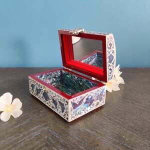 May include: An open, rectangular jewelry box with a red interior and a mirrored lid. The box is decorated with a white and blue floral pattern. The interior base has an iridescent, abalone-like finish. A small silver clasp secures the lid.