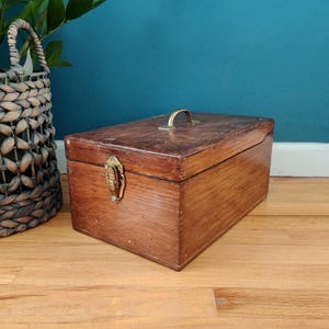 May include: A rectangular wooden storage box with a medium brown finish, featuring a brass-colored metal handle on the top lid and a matching brass-colored metal latch on the front. The box has a visible wood grain texture and a hinged lid design.