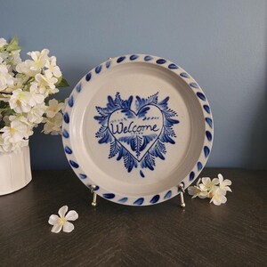 May include: A round, off-white ceramic plate featuring a hand-painted blue heart design in the center with the word Welcome written inside. The rim of the plate is decorated with a repeating pattern of small blue leaf-like shapes around the edge.