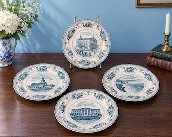May include: A set of four round ceramic plates featuring blue and white transferware patterns. Each plate displays a different architectural scene in the center, surrounded by a decorative floral border with scalloped edges.