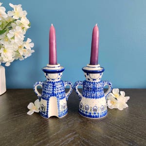 May include: Two ceramic candle holders with a blue and white pattern, each holding a purple candle. The candle holders have a whimsical design with a face and arms. The background is a solid blue.