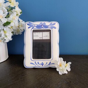 May include: White ceramic picture frame with blue floral and bird design. The frame has a rectangular opening and a grid pattern inside. The frame is 9 x 13 inches.