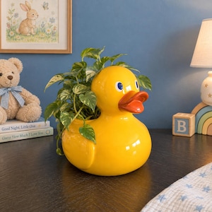 May include: A bright yellow ceramic duck-shaped planter featuring a glossy finish, a vibrant orange beak, and blue eyes. The duck is filled with a lush green and white variegated pothos plant, creating a cheerful and functional decorative piece.
