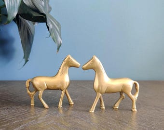 May include: Two gold-colored horse figurines. The horses are facing each other, with their heads nearly touching. The figurines are on a wooden surface, with a blue wall in the background. A green plant is visible in the upper left corner.