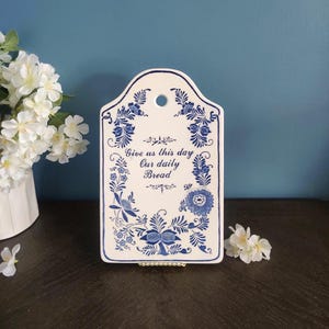 May include: White ceramic bread board with blue floral designs and the text "Give us this day Our daily Bread." The board has a hole at the top for hanging and a decorative border. A charming addition to any kitchen.