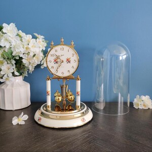 May include: A gold and white anniversary clock with a floral pattern on the face, base, and pillars. The clock features a rotating pendulum with four gold spheres, a white dial with black numerals, and a clear glass dome cover.