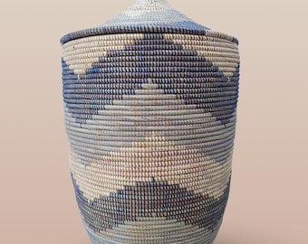 May include: A woven storage basket with a lid. The basket is cylindrical with a rounded base and a matching lid with a small knob. The basket features a pattern of blue, white, and tan horizontal stripes and angled shapes.