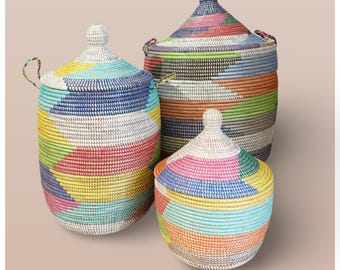 May include: Three colorful woven baskets with lids. The baskets are made of natural materials and have a geometric pattern. The largest basket is green, blue, and white. The medium basket is pink, blue, and yellow. The smallest basket is pink, blue, and green.