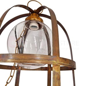 May include: Close-up of a vintage-style pendant light fixture. The fixture features a clear glass globe encased in a cage of aged brass-colored metal. A chain is attached to the globe. The light fixture is designed to be suspended from the ceiling.