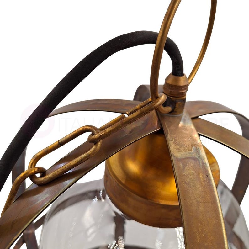May include: Close-up of a vintage-style pendant light fixture. The fixture features a clear glass globe encased in a cage of aged brass-colored metal. A black electrical cord and chain are visible, adding to the industrial aesthetic. The light fixture is designed for ceiling mounting.