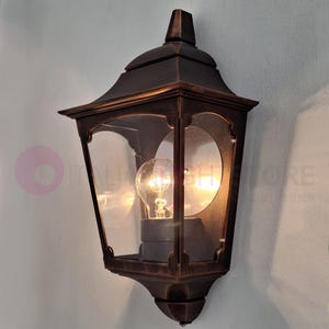 Könnte beinhalten: Eine bronzefarbene Außenwandleuchte mit einem klaren Glasschirm und zwei Glühbirnen. Die Leuchte hat ein traditionelles Design mit einem dekorativen Oberteil und einem quadratischen Sockel.